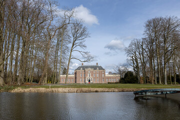 Fototapeta na wymiar Painterly Dutch landscape with exterior facade of Slot Zeist castle behind winter barren trees on a sunny day with park surrounding it and moat in the foreground against a blue sky with fluffy clouds