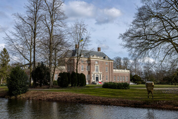 Fototapeta premium Sideview of exterior facade of Slot Zeist castle behind winter barren trees on a sunny day with park surrounding it and moat in the foreground against a clue sky with clouds
