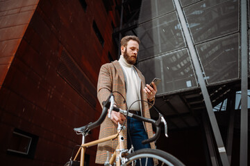 Young man with bicycle holding a smartphone front of building. Creative businessman in a modern business area.