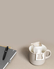 Drip coffee bag with ground coffee in big cup on office table, package with paper bag drip coffee filter for brewing on beige color background, side view with copy space,