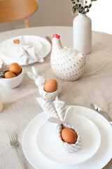 Fototapeta na wymiar Easter egg with rabbit ears made of napkin on plate, cutlery, decor on desk with linen tablecloth.