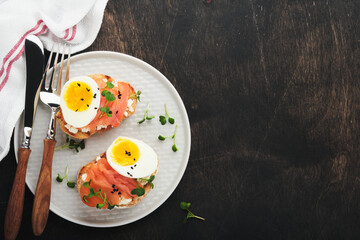 Sandwich with delicious toppings smoked salmon, eggs, herbs and microgreens radish, black sesame seeds over white plate on old wooden table background. Healthy open sandwich superfood. Top view.