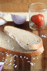 Sliced bread lies on a wooden board. Nearby are onions and pickled tomatoes. Coarse salt is scattered on the board.