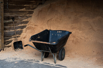 Big pile of sawdust with empty wheelbarrow and a shovel in horse stable on ranch