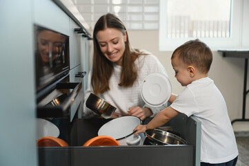 Happy mom and cheerful baby in the kitchen. A restless child sorts through the dishes in the...