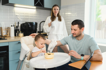 Young parents father and mother feeding the baby. Child on a highchair in the kitchen. Dad, mom and baby - happy full family