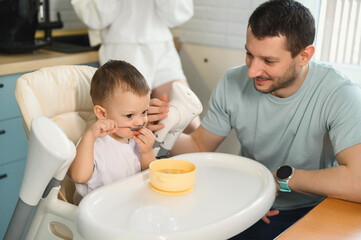 Young parents father and mother feeding the baby. Child on a highchair in the kitchen. Dad, mom and baby - happy full family