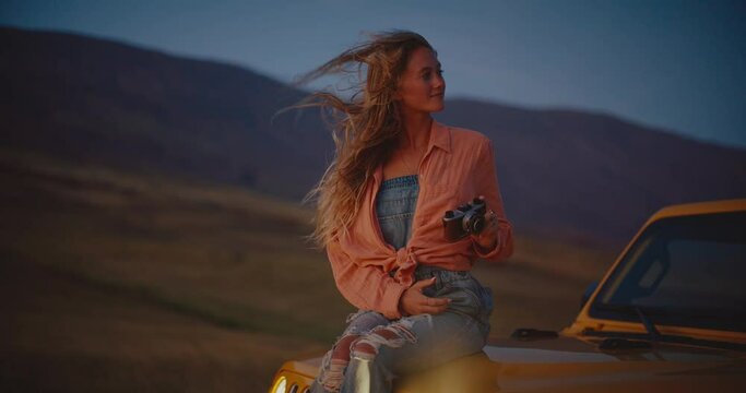 Beautiful young woman taking pictures at dusk with vintage camera, hair blowing in the wind in slow motion, travel and adventure nomadic lifestyle