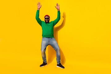 Fototapeta na wymiar Photo of positive man dance raise hands wear sunglass red beanie green shirt jeans isolated yellow color background