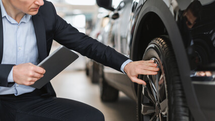 Unrecognizable auto salesman sitting near brand new car, checking wheel and tire at modern dealership, closeup