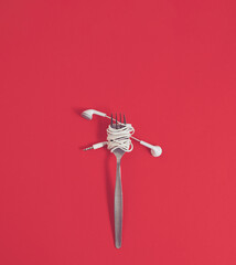 Earphones wrapped around a fork like spaghetti. Vertical flat lay compsition on a pastel red basground, minimal music enjoyment concept