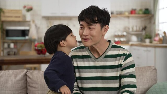 slow motion of Asian baby boy feeling shy after kissing dad on face in the living room at home. happy father embracing his son who is looking down with embarrassment