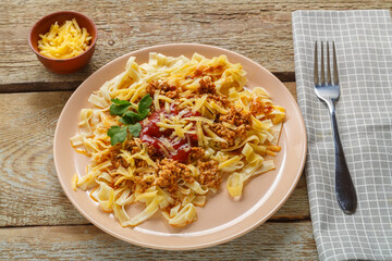 Pasta bolognese in tomato garnished with herbs and cheese in a plate on a wooden table next to a fork and napkin and grated cheese in a plate.