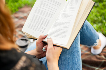 A woman in jeans with cup of coffee reading book while sitting on a blanket in the park