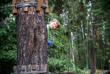 Strong excited young boy playing outdoors in rope park. Caucasian child dressed in casual clothes and sneakers at warm sunny day. Active leisure time with children concept