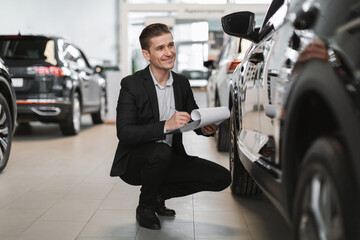 Fototapeta na wymiar Happy young car salesman sitting near new auto, checking it, taking notes at automobile dealership