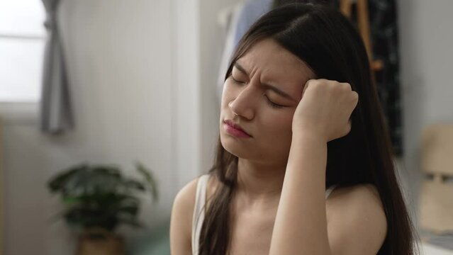 headshot of dizzy asian female massaging herself with grimacing face while trying to ease pain in the head caused by hangover after rising in the morning at home