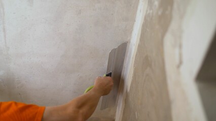 Master is applying white putty on a wall and smearing by putty knife in a room of renovating house in daytime. The worker applies the putty to the wall with a putty knife.