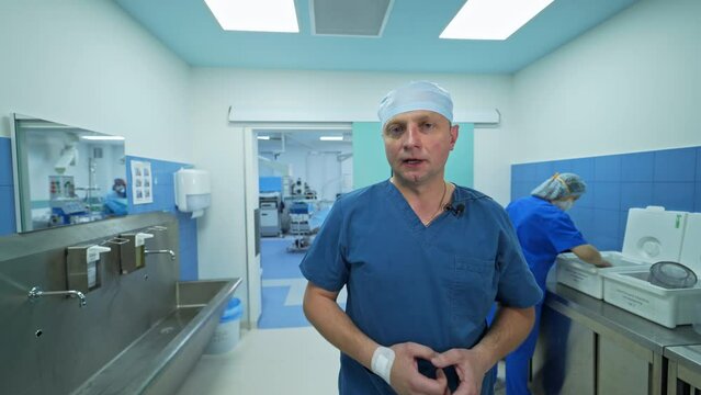 Experienced male surgeon talks to the camera standing in the preoperational room. Female nurse dealing with instruments at backdrop.
