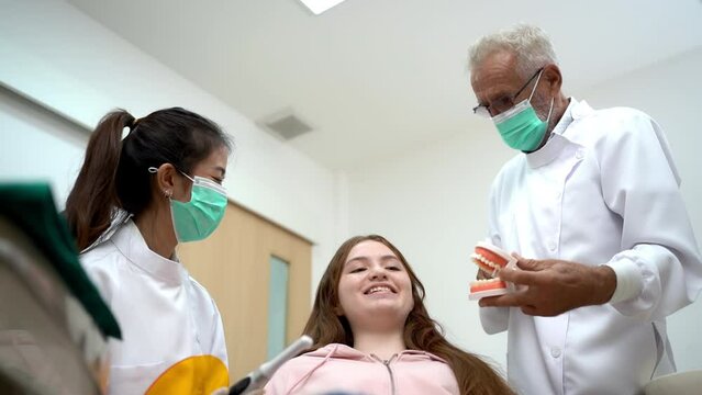 Senior Caucasian male dentist in a face mask explaining the procedure of dental treatment using teeth model to a Caucasian teenage girl before doing a dental work by him and a young Asian female nurse