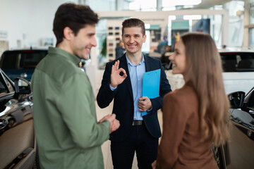 Auto showroom manager showing okay gesture to happy young customers at car dealership