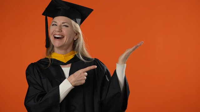 Portrait of happy education graduate student woman in mortar board with diploma laughing finger pointing to advertising on orange background