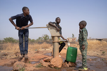 Three African children filling up water in a plastic tank at a borehole pump in a rural community in Sub-Saharan Africa. Child labor concept. - obrazy, fototapety, plakaty