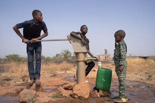 Three African Children Filling Up Water In A Plastic Tank At A Borehole Pump In A Rural Community In Sub-Saharan Africa. Child Labor Concept.