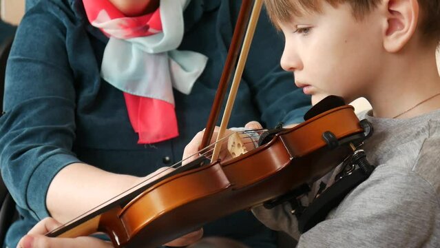 A woman teaches a boy to play the violin holding his right hand in her hand and leading the bow along the string