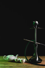 Tea in cups and teapot near hookah on wooden surface isolated on black.