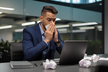 Sick middle eastern businessman sitting in his office, sneezing
