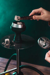 Cropped view of man holding coal in tweezers near hookah on green background with smoke. © LIGHTFIELD STUDIOS