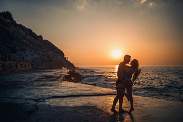 Fototapeta premium Beautiful couple in love on the background of the sunset by the sea. Young woman and man hugging by the sea at sunset