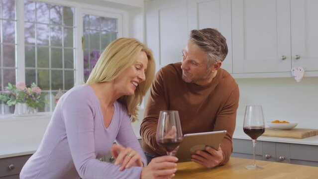 Mature couple at home in kitchen using digital tablet to book show, vacation or make purchase and celebrating by drinking red wine together - shot in slow motion