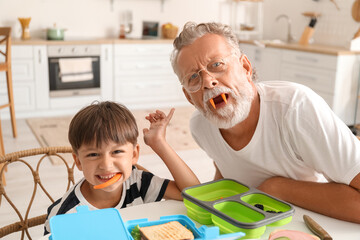 Little boy with his grandfather having fun while packing lunch boxes at table in kitchen