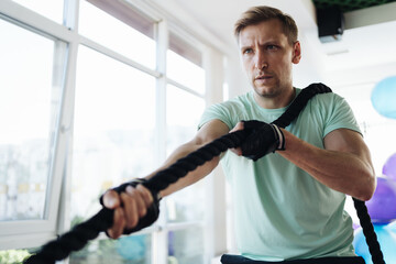 Caucasian blonde man training with rope in functional training fitness gym with big windows and a lot of daylight.