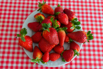 Fresh strawberries with green leaves in a white plate on a red and white tablecloth.