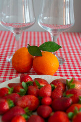 Two oranges with green leaves on a red and white tablecloth and glass wine glasses.