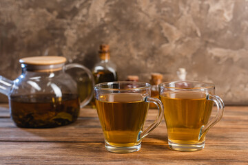 Cups of tea near blurred teapot on wooden surface on textured stone background.