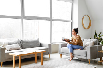 Beautiful young woman reading magazine on couch at home