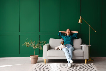 Young woman with Jack Russel terrier resting on grey sofa near green wall