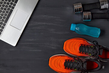 Orange sneakers, dumbbells, a water bottle and a laptop on a dark wooden background