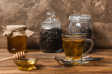 Cup of tea near honey and blurred jars on wooden surface on textured stone background.