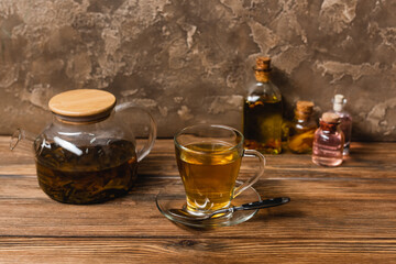 Cup of tea near teapot and blurred bottles on wooden surface on textured stone background.