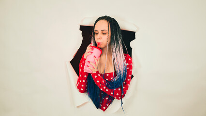 Barmaid with drink looking through hole in paper background. Female bartender with dreadlocks and cup of sweet beverage peeking through ripped white paper background in studio and looking at camera