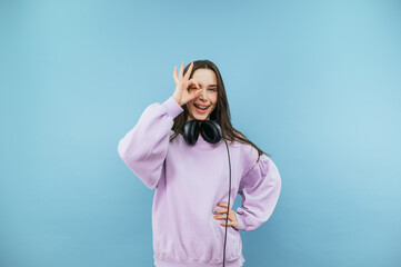Funny girl in a sweatshirt and headphones stands on a blue background and shows a gesture ok winking at the camera