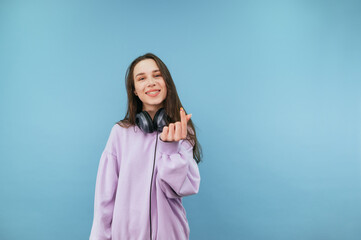 Fototapeta premium Positive girl in headphones stands on a blue background in headphones and shows a heart gesture to the camera with a smile on his face.