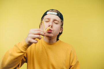 Hungry man in cap and eating french fries with closed eyes on yellow background, close up photo....