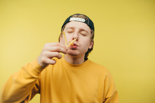 Hungry Man In Cap And Eating French Fries With Closed Eyes On Yellow Background, Close Up Photo. Guy And Fast Food.