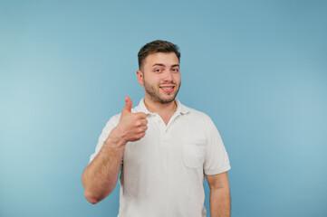 Positive young man with bristles in a white T-shirt stands on a blue background and shows a gesture...